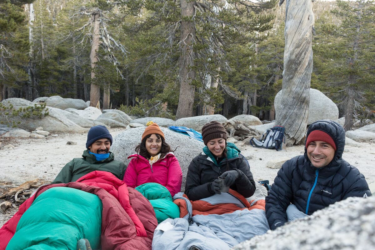 Four people sitting side-by-side bundled up in warm gear and sleeping bags on a cold weather camping trip