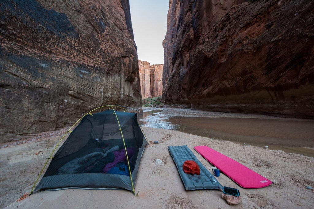 Tent and sleeping pads set up on sandy beach in Canyon with river running through it and tall canyon walls on either side