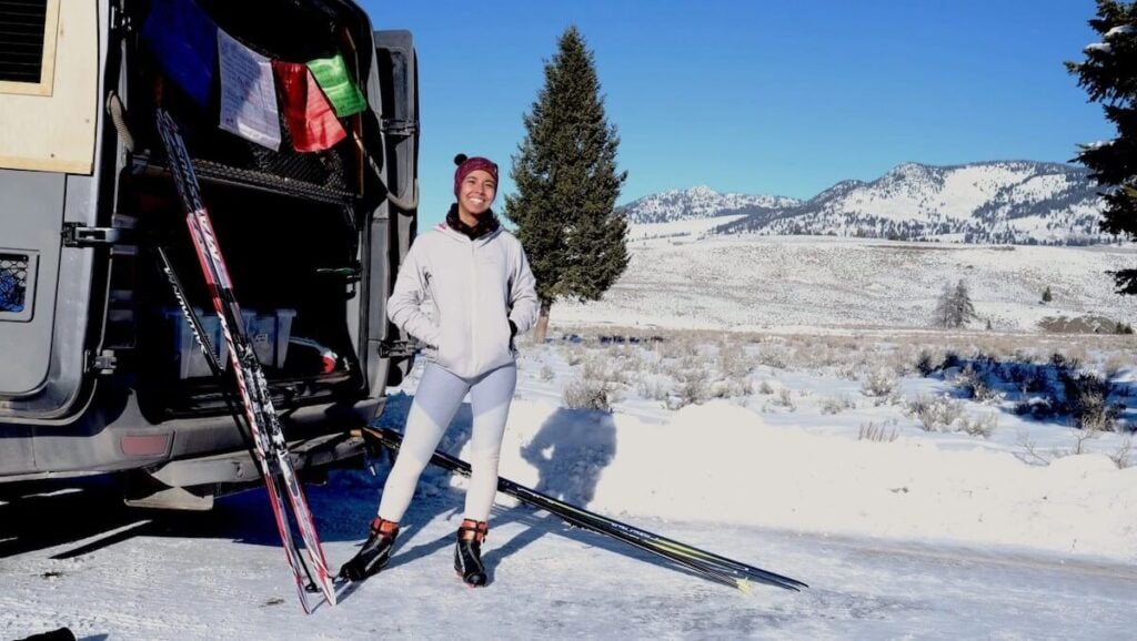 a woman standing at the back of her van with the doors open, ski gear leaned against the vehicle.