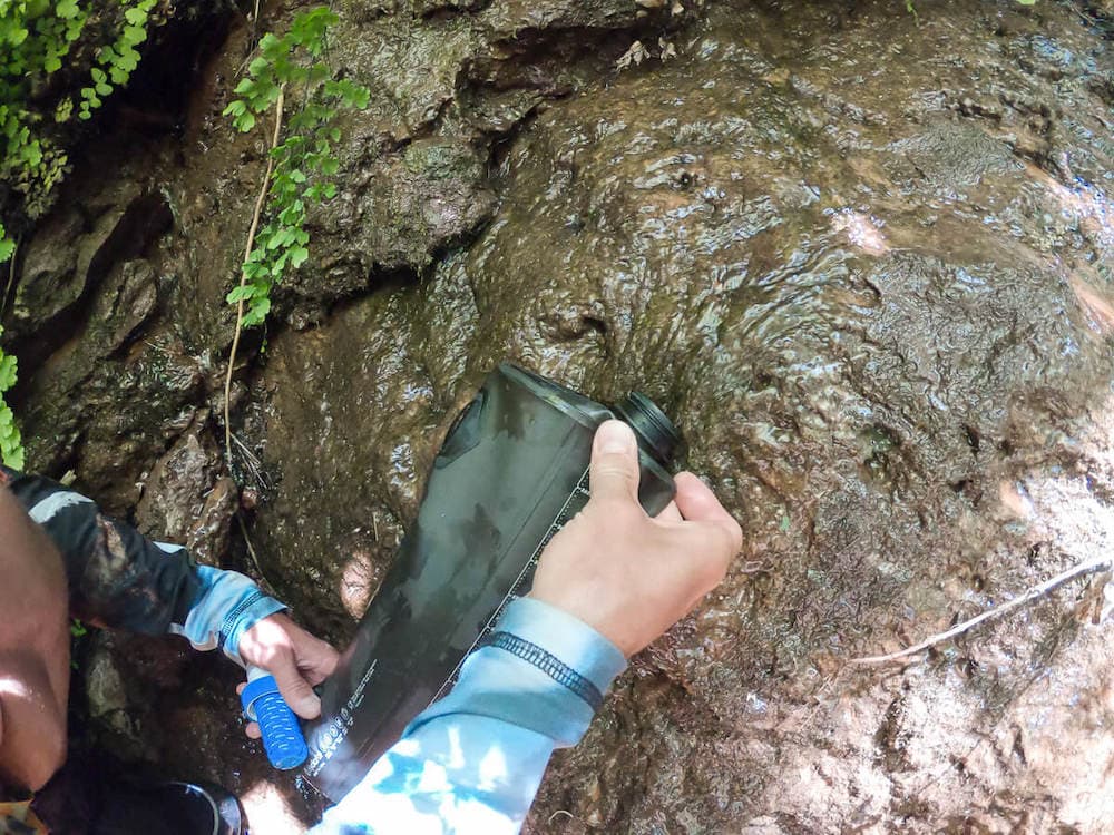 Person filling water filter reservoir from shallow, muddy puddle