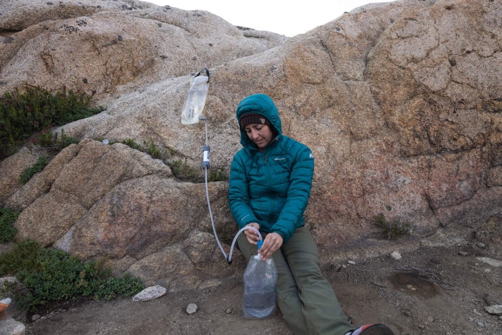 Woman sitting with back against large rock using gravity-fed water filter to filter drinking water. 