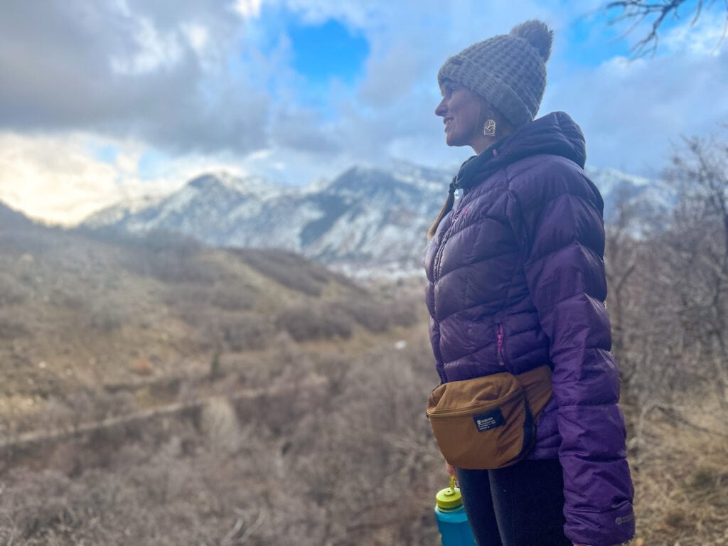 Woman wearing lululmeon fanny pack and warm puffy jacket on hike with snowcapped mountains in background