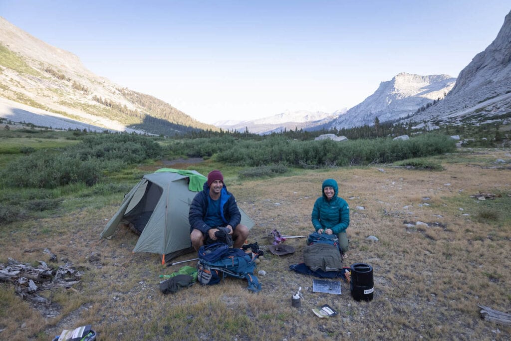 Two backpackers sitting on ground at camp with backpacking gear laid out around them and mountains in background