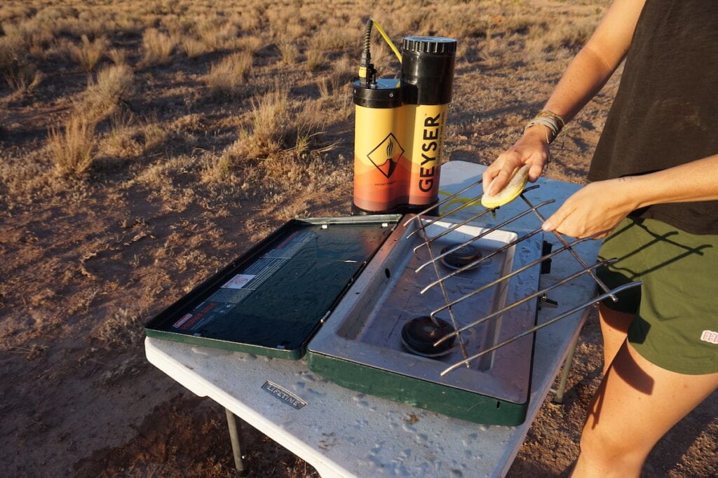 A woman scrubs her camp stove using the Geyser Portable Shower