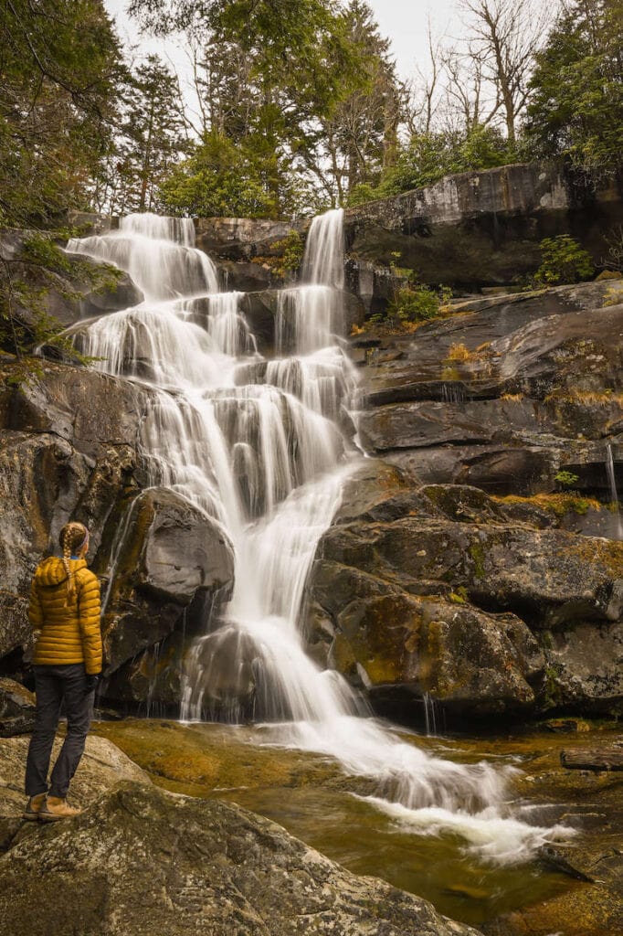 Woman standing on rocks at base of waterfall looking up at cascade