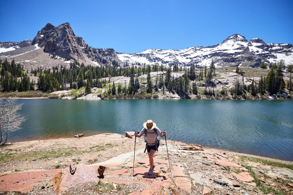 A woman hikes up a rocky hill next to  a lake using trekking poles