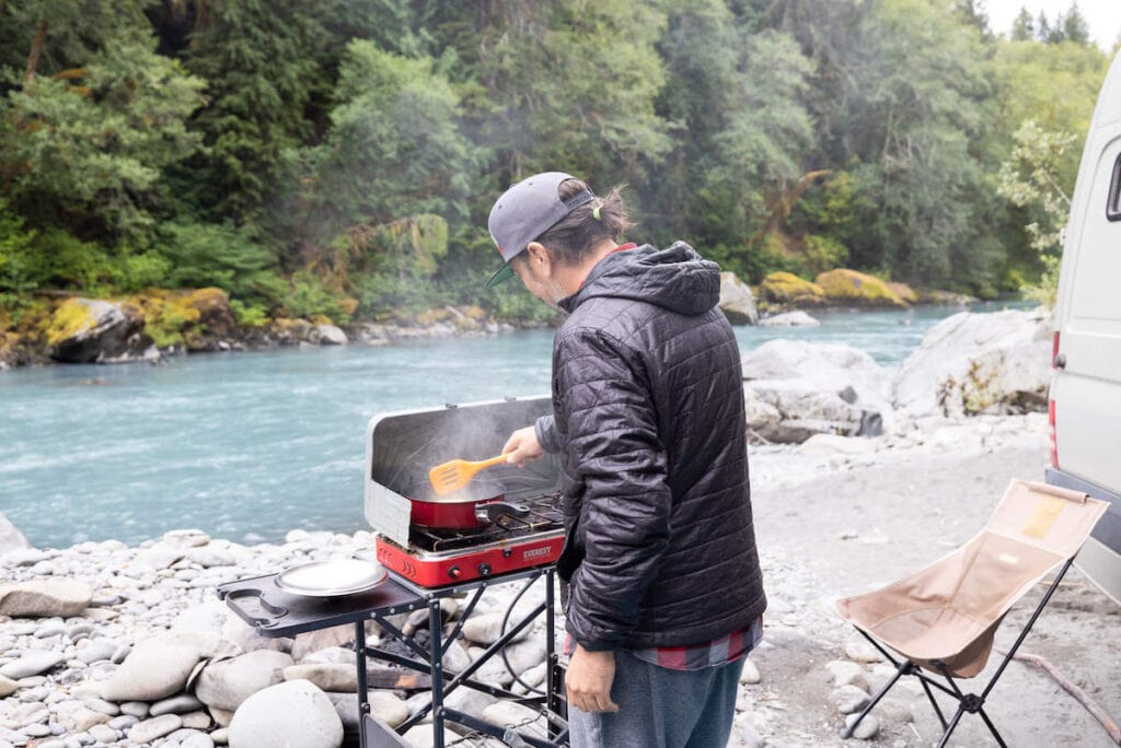 Man cooking on outdoor stove in front of river at campsite