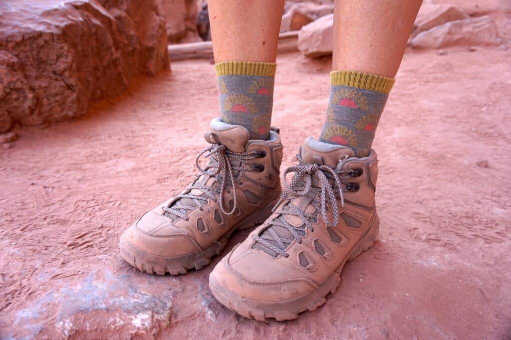 A close up of a woman wearing Oboz Sawtooth X Mid Hiking Boots and Darn Tough hiking socks. There is red rock surrounding her