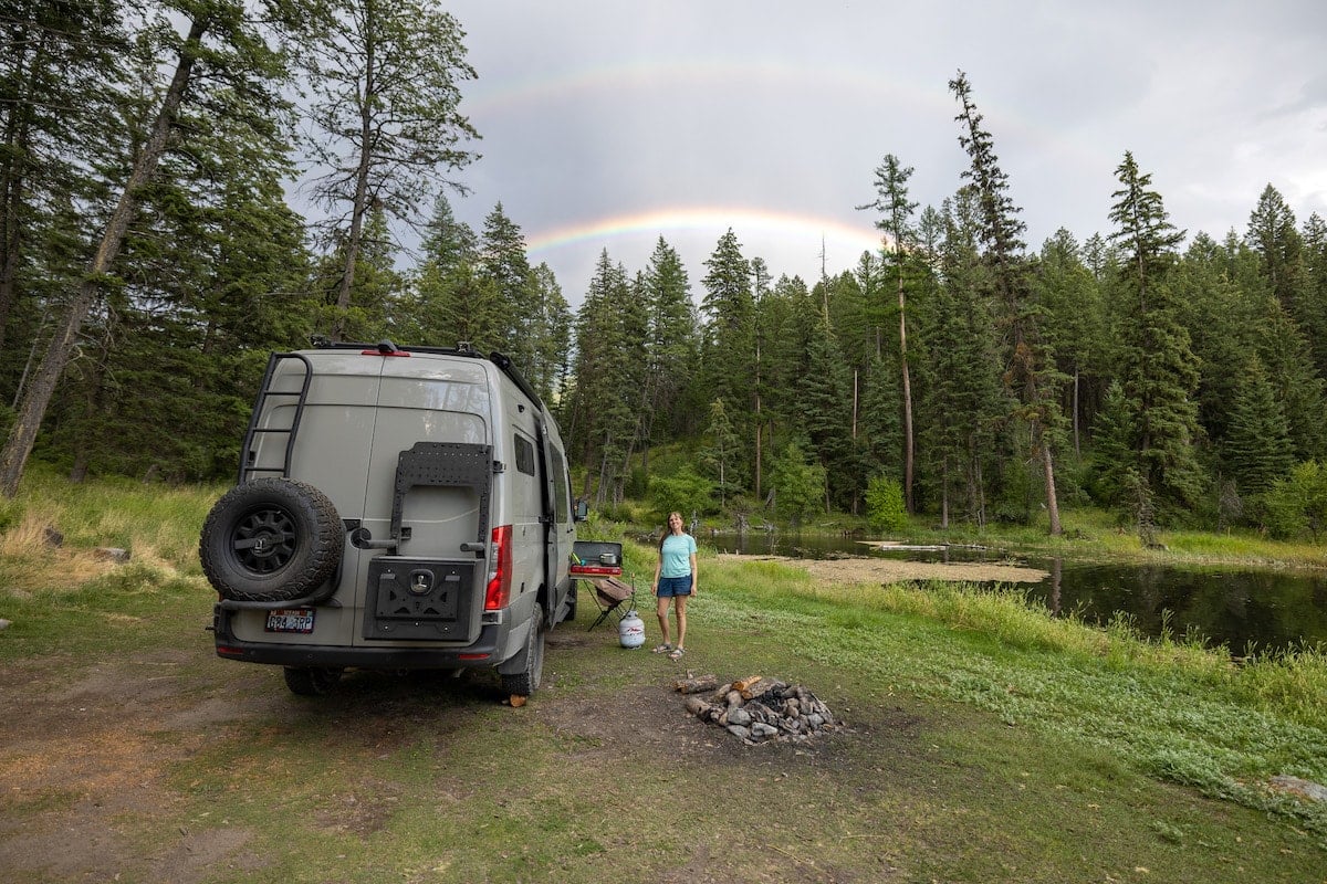 A woman stands under a rainbow with her Sprinter van. The Owl Vans B2 carrier, Mini-Sherpa mounting plate, and medium expedition box are on our right rear door of her Sprinter Van