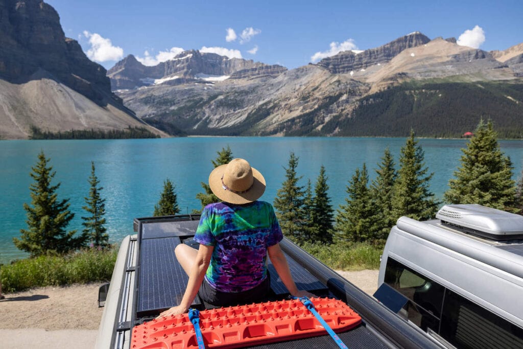 Woman sitting on top of converted Sprinter van looking out over blue water of Bow Lake in Canadian Rockies