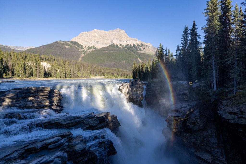 Athabasca Falls in Alberta with rainbow on one side and towering mountain peak in distance