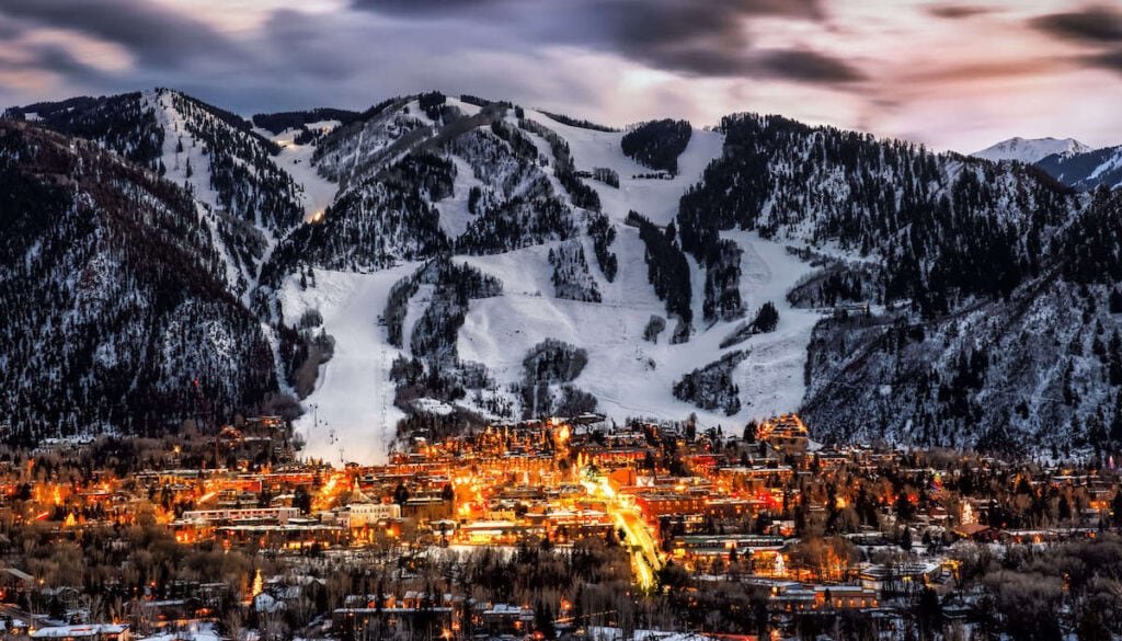 Views of Aspen Colorado at dusk with lights illuminating the town and ski slopes running straight down into Aspen