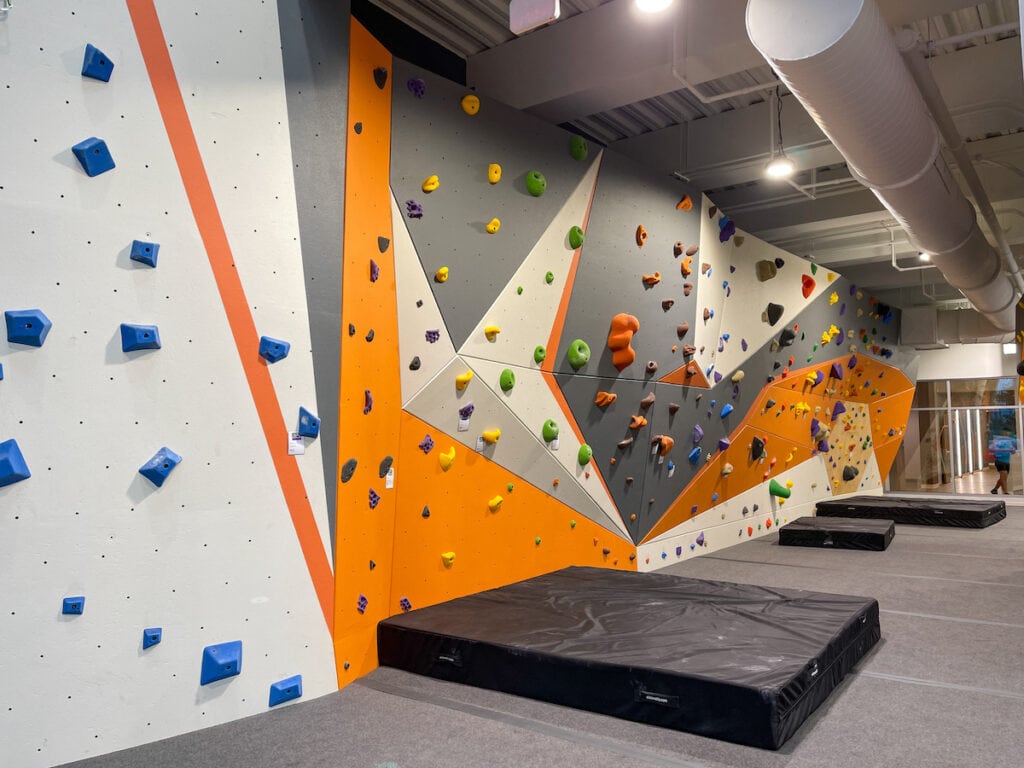 An landscape shot of an empty bouldering wall at a rock climbing gym with black crash pads under routes