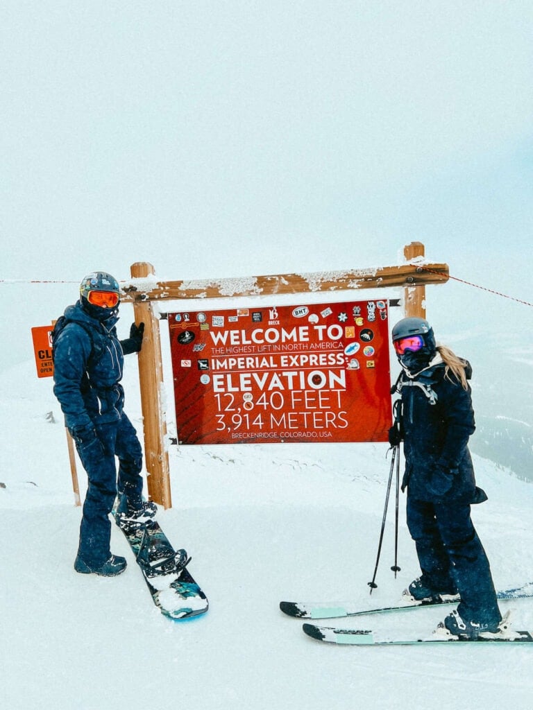 Skier and snowboarder standing next to sign at Breckenridge Resort that says "welcome to the highest lift in North America: Imperial Express. Elevation 12,840 feet, 3,914 meters"