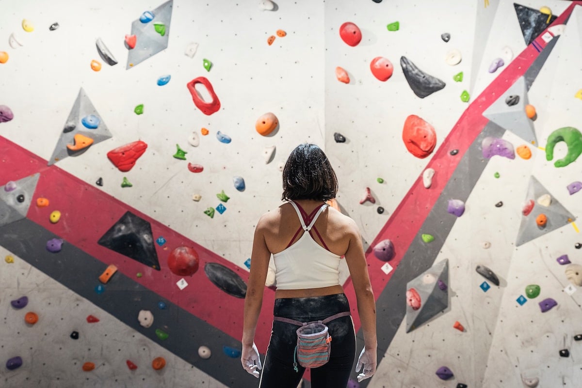 A woman stands on the ground facing a bouldering wall while indoor rock climbing