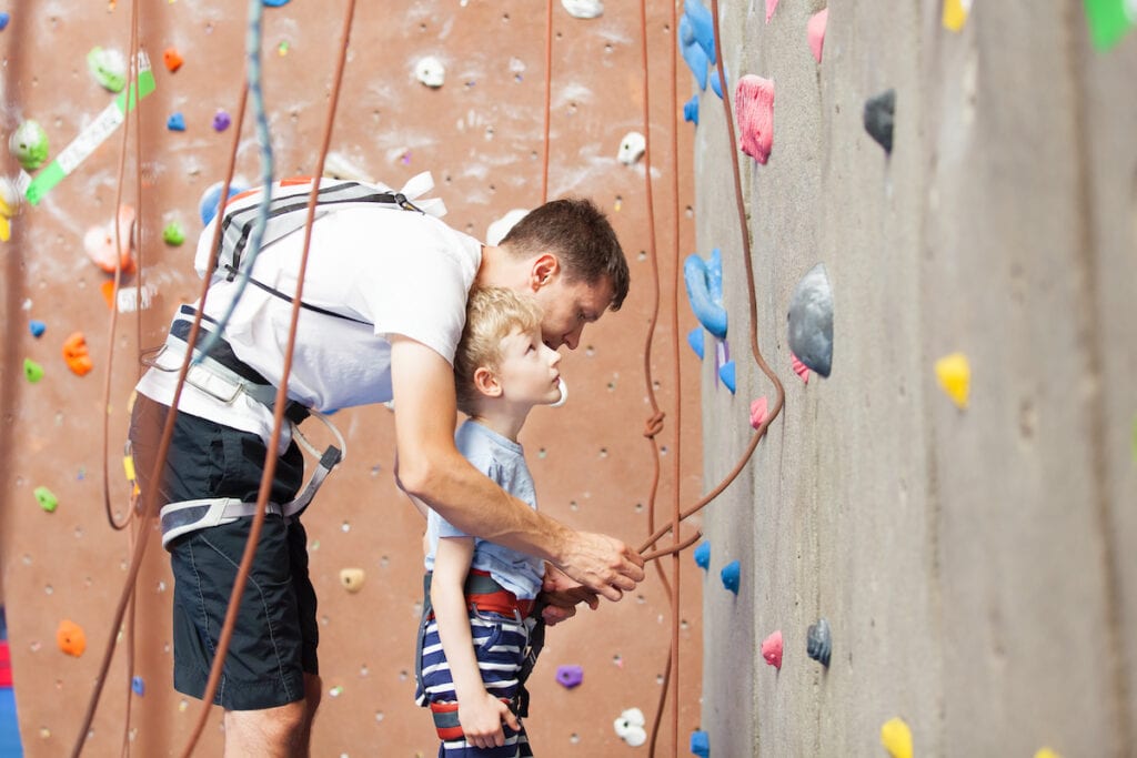 A father hooks his son into a belay device before starting a climb at an indoor rocking climbing gym. The boy is looking at the climbing wall.