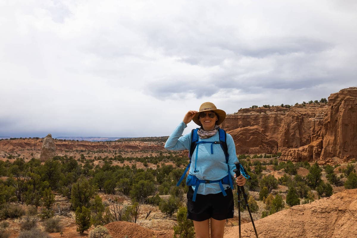 Hiker smiling for camera wearing hiking gear and carrying trekking poles on trail in Utah with red rock bluffs in background