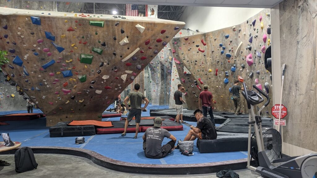 A landscape image of an indoor bouldering area of a climbing gym. There are four men assessing the wall and two men sitting on the ground nearby watching.