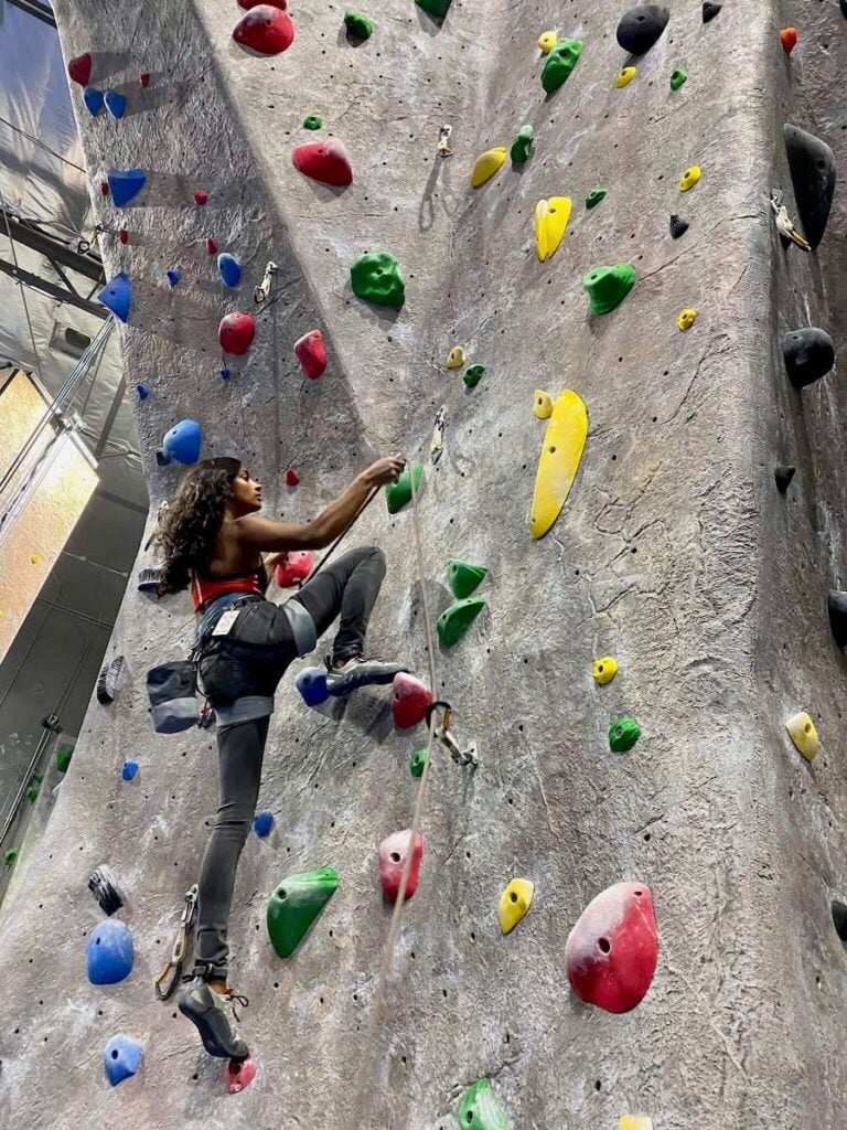 A woman climbing at an indoor rock climbing gym wearing a tank top and leggings