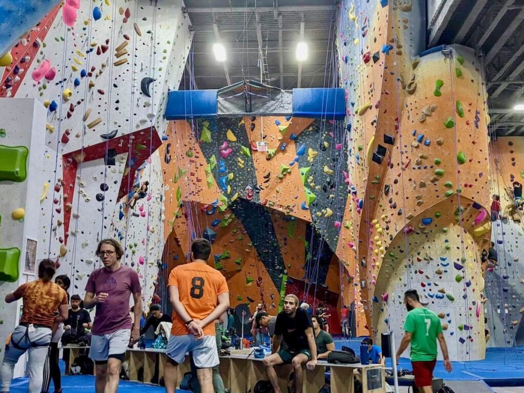 A landscape shot of an indoor climbing gym - many people sitting around and watching climbers on the wall