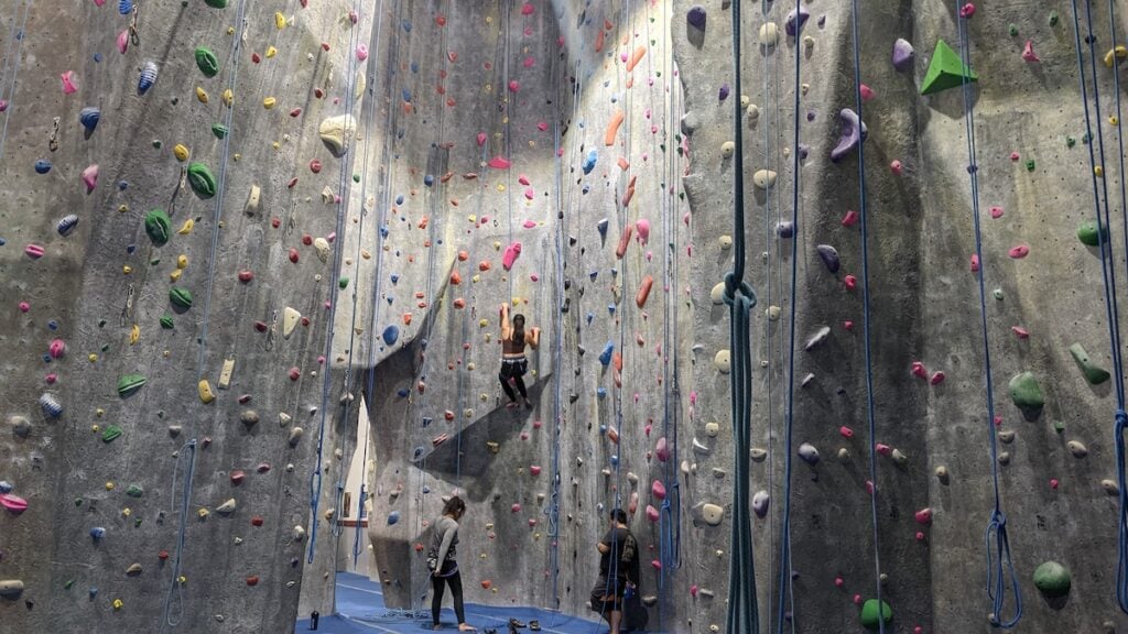 A woman rock climbs in an indoor climbing gym using a top rope with a belayer under her