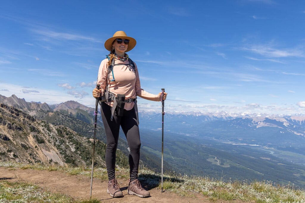 Kristen hiking in front of a mountainous background wearing a hat, REI Sahara Shade Hoody, leggings, and Oboz Sypes hiking boots, some of the best gifts for outdoor women