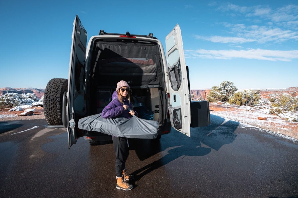 A woman smiles at the camera while holding a YETI Trailhead Camp Chair while carrying a bag over her shoulder.  She's standing next to the Sprinter with the back doors open