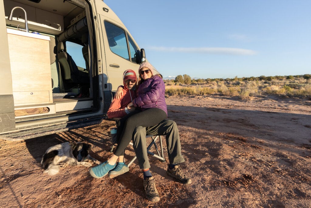 A man sits on a YETI Trailhead Camp chair with a woman on his lap.  Sprinter camper van in the background and dog lying beside chair.