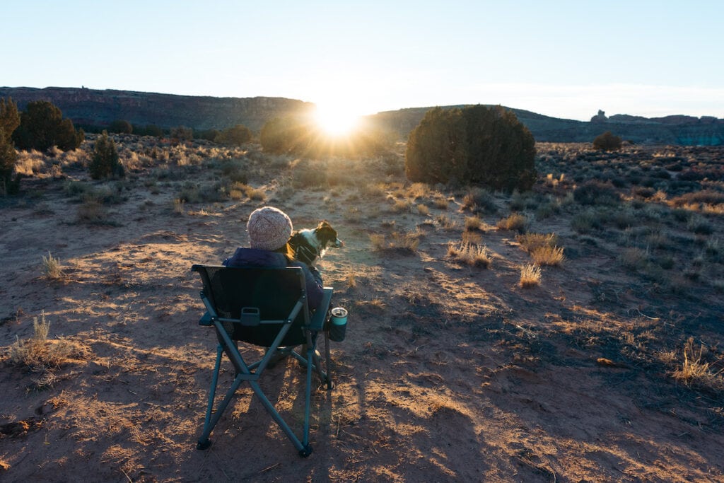 A woman sits on a YETI Trailhead Camp chair with a dog on her lap as the sun sets in front of them in Moab, Utah