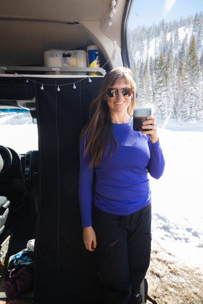 A woman holds a cup of coffee standing in the side door of her Sprinter Van wearing Icebreaker 200 Oasis Crew Top.