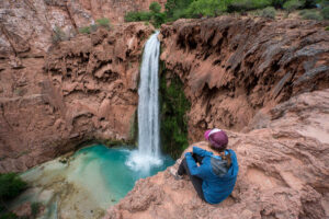 Woman sitting on rock ledge looking down over Mooney Falls in Havasupai with turquoise water and red rock cliffs