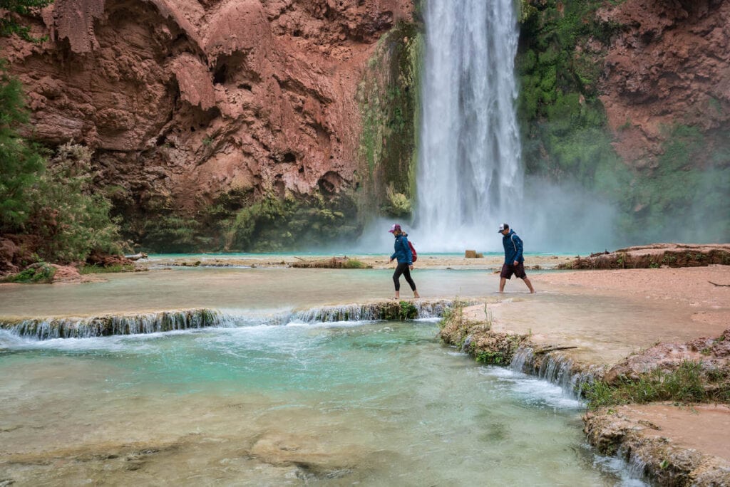 Two people walking across turquoise water at base of Havasu Falls in Arizona