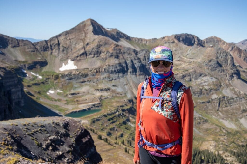 Kristen smiling for photo on hike Colorado wearing hat, buff, sunglasses, and long sleeve shirt