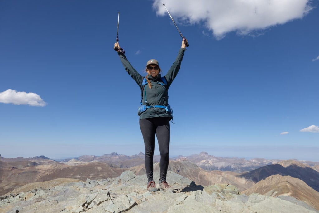 Woman standing on rocky Handies Peak summit wearing hiking gear and arms raised above her head
