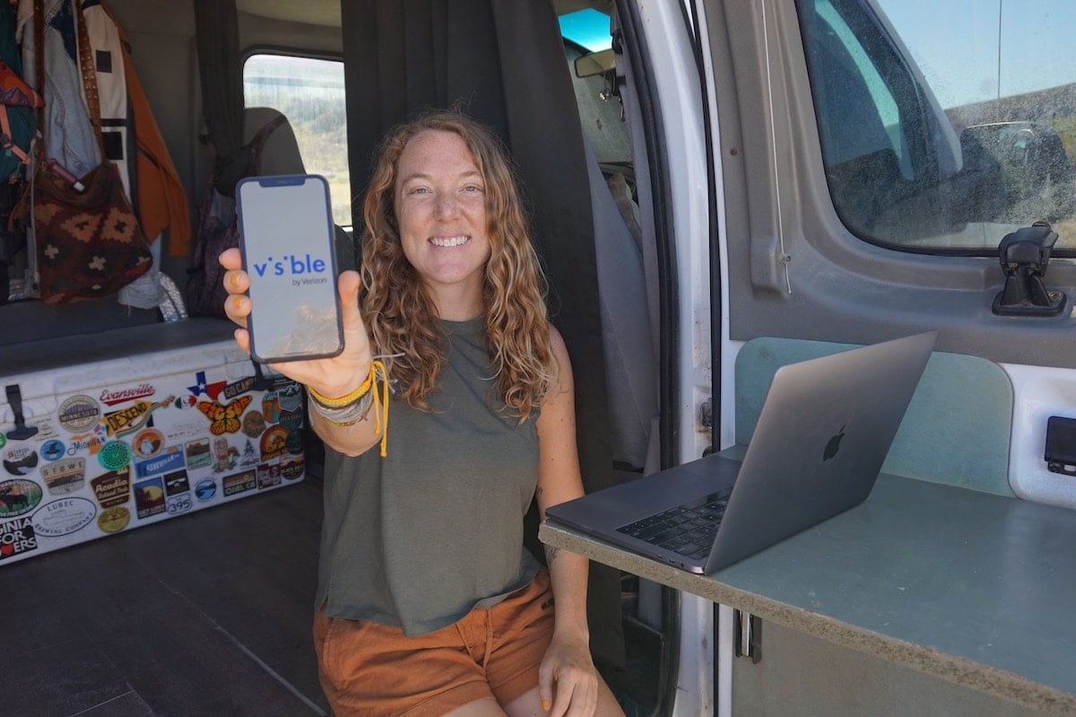 A woman sits in the doorway of her van holding a phone with Visible Wireless logo on the screen and a laptop on a table nearby