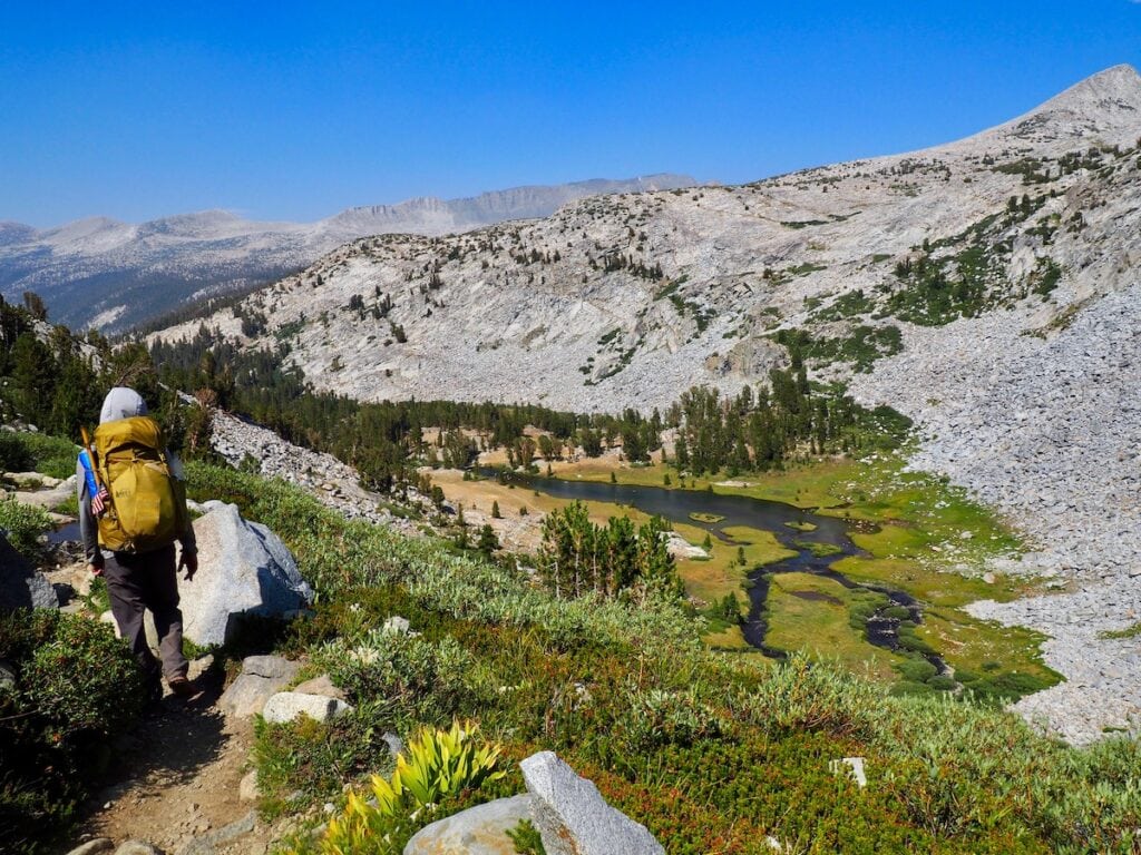 A man stops to view Lyell Pass on the John Muir Trail. There are mountains and a creek in the distance