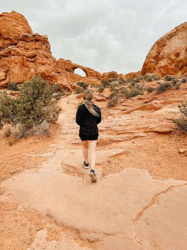 Woman hiking on red rock trail in Arches National Park with rock arch in distance