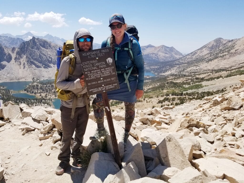 A man and woman smile next to a hiking sign "Kearsarge Pass: 11,760 feet. Entering Kings Canyon National Park" on the John Muir Trail