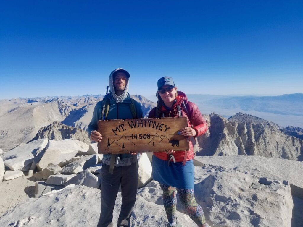 A man and woman smiling holding a "Mt Whitney 14,508ft" sign 