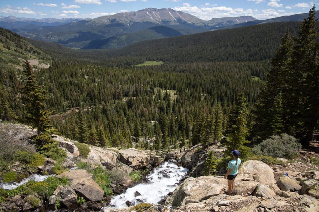 Woman hiker standing alongside a creek looking out at the views of a forested landscape below with mountains in the distance while hiking in Breckenridge