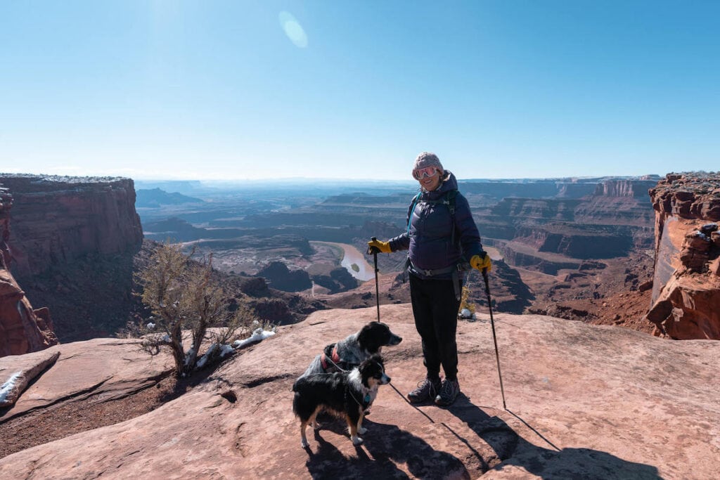 Woman with hiking poles and dogs standing at Dead Horse State Park lookout over Colorado River and Canyonlands National Park in Utah