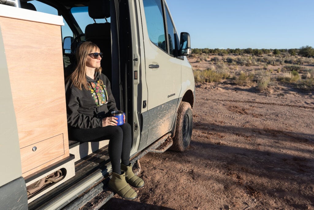 A woman holds a YETI Rambler Mug sitting in the doorway of her Sprinter van. She has REI Co-op Camp Dreamer Booties on her feet.