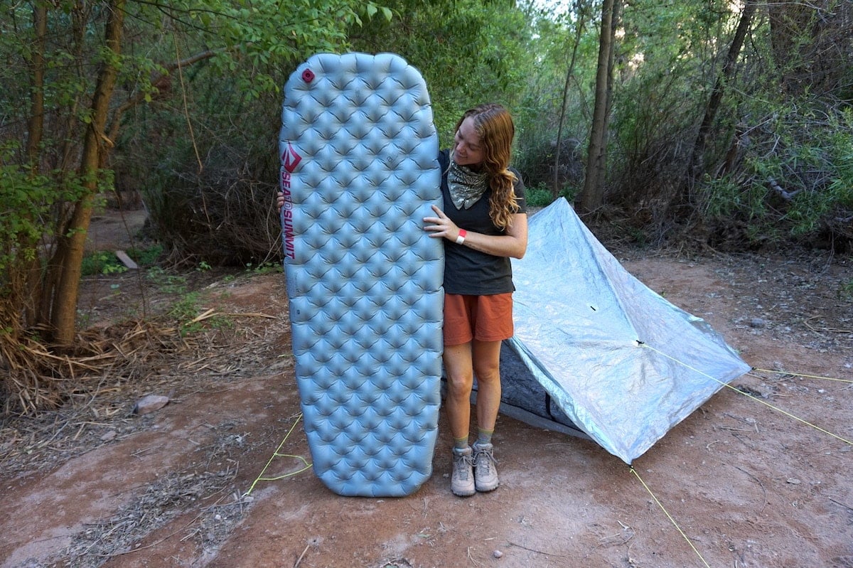 a woman stands next to an inflated Sea to Summit Ether Lite Sleeping Pad with a backpacking tent set up in the background