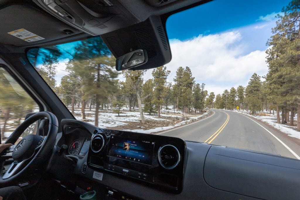 View out the front windshield of the Storyteller Overland Classic MODE AWD van showing the front cab, touchscreen display, and a snowy forested road