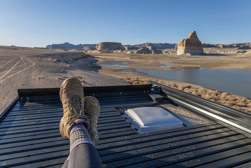 Woman sitting on the roof rack on top of the Storyteller Overland Classic MODE van with Lone Rock at Lake Powell in the background