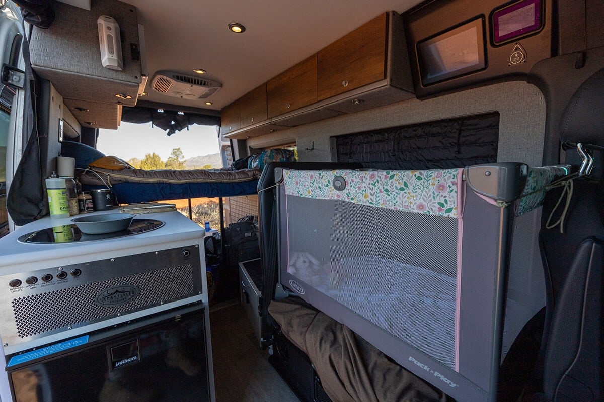 Interior of the Storyteller Overland Classic MODE van showing the sleeping area with a Pack n Play set up for a toddler