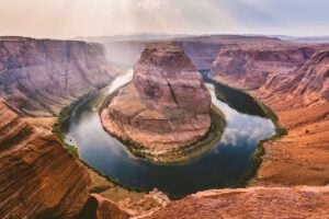 Turquoise waters of the Colorado River flow in a sweeping arc at the base of towering rust-colored cliffs. The view from above showcases Horseshoe Bend in Arizona, where the river makes a dramatic 180-degree turn, surrounded by layers of eroded rock formations. The sunlight casts a warm glow on the landscape, highlighting the textures and curves of the rock formations.