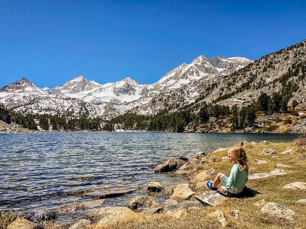 Little girl sitting by edge of lake with towering snow-capped mountains rising above