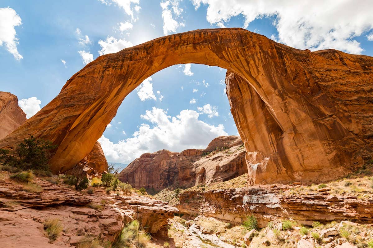 A massive natural rock arch rises up against a clear blue sky at Rainbow Bridge National Monument. The majestic Rainbow Bridge spans 275 feet across, towering over the surrounding terrain. The sandstone formation has been shaped over millions of years by the forces of wind and water, creating a breathtaking natural wonder. The colors of the rock range from warm reds and oranges to cool grays and blues, adding to the natural beauty of the scene.