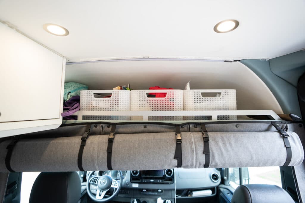 Camper van organization ideas showing three bins on the overhead shelf above the cab in a Sprinter Van